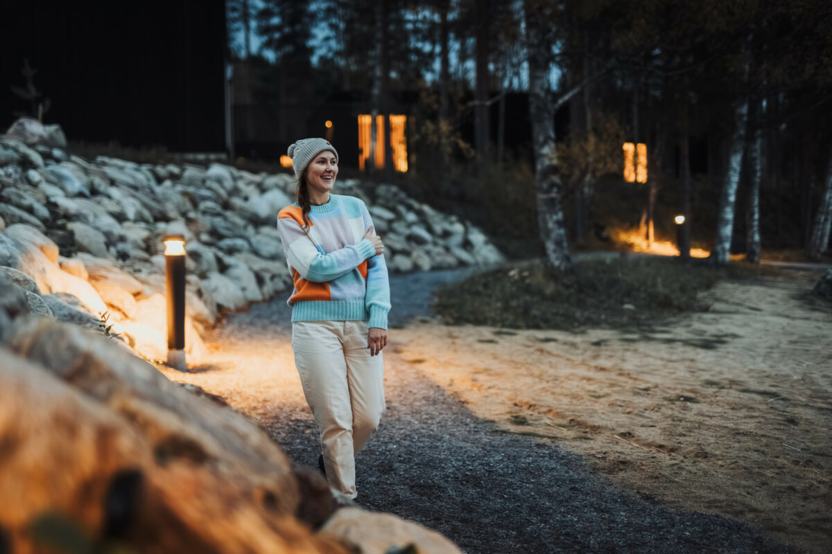 Naurava nuori nainen kävelee hämärtyvässä syysillassa hiekkarannalla. Taustalla näkyy tummia puurakenteisia rakennuksia, joiden ikkunoista loistaa valoa.