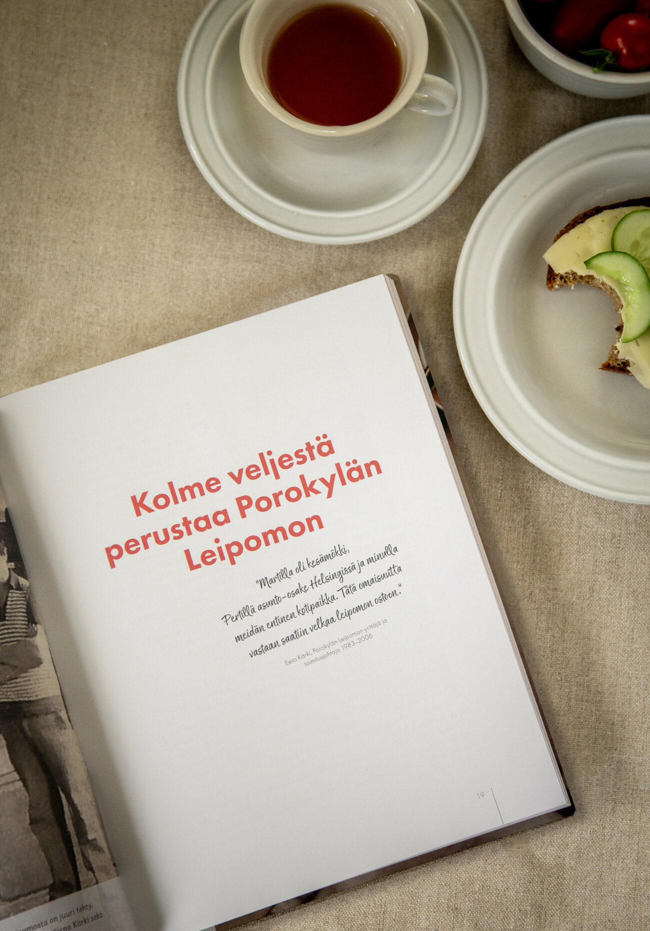 Porokylän Leipomon historiikkikirja avattuna aukeamalta jossa näkyy otsikkona Kolme veljestä perustaa Porokylän Leipomon. Kirja on aseteltu pellavaliinalle ja liinan vieressä on teekuppi ja lautanen jossa on osittain syöty ruisleipä.